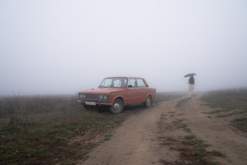 Red retro car stands near the dirt road in fog, young girl in white coat stands under umbrella