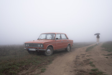 Old red retro car stands in fog near the dirt road, girl in white coat walks under umbrella on the background