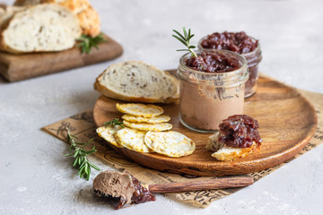 Fresh homemade chicken liver pate and onion chutney (marmalade) in a jar, bread and crackers on a wooden plate. Light grey background, selective focus.