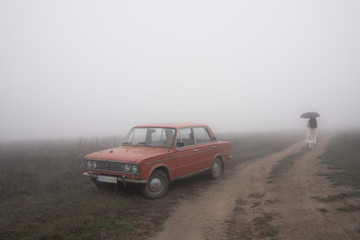 Fototapeta na wymiar Old red retro car stands in fog near the dirt road, girl in white coat walks under umbrella on the background