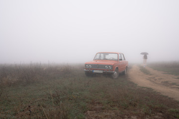 Fototapeta na wymiar Red old retro car near the dirt road, young girl in white coat under umbrella walks in the fog on the background