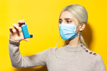 Studio portrait of young blonde girl, wearing medical flu mask and beige sweater, holding asthmatic inhaler in hand on yellow background. - obrazy, fototapety, plakaty