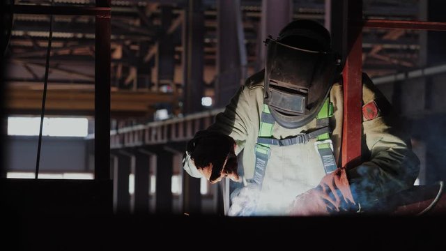 Industrial worker in protective mask and safety gloves welds metal structures on construction site. Worker in face shield uses gas welding torch. Fire flickers and glowes, orange sparks are flying. 