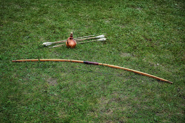 Ancient ceramic jug, wooden long bow and arrows lying on the ground with green grass. Historical reenactment concept, hobbies and leisure