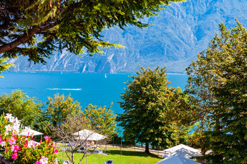 Walkway near the coast of Garda lake with blooming flowers, high evergreen trees, green lawns full of open summer tents, resting tourists and gorgeous lake with dolomite mountains on background