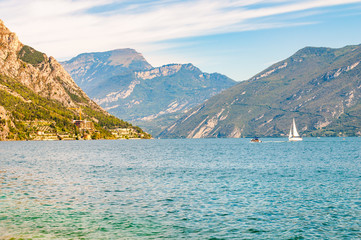 Beautiful Garda lake in Italy surrounded by high dolomite mountains. Classic white sailing yacht floating on the lake. Various hotels and private houses built on the rocky shores of the lake