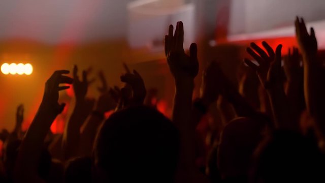 Super slow motion: people crowd partying, cheering, raising hands up and clapping at rock concert in front of stage of nightclub. Bright colorful stage lighting. Nightlife and entertainment concept