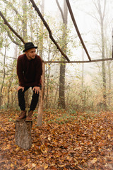 Attractive young man in red sweater and black hat crouched on the stump under the dry log canopy among the forest in fog