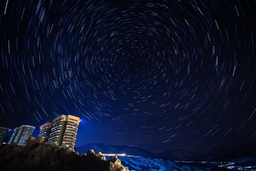 Fototapeta premium Star trail panorama over the apartment blocks in the ski village of Le Corbier, high in the French Alps around midnight. Star Polaris is seen in the center.