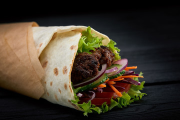 Shawarma rolled in lavash, moist grilled meat with onion, herbs and vegetables on wooden black background.