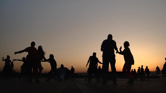 Unrecognizable people silhouettes learning how to dance on city quay at sunset - super slow motion. Warm illumination, evening time. Romantic pairs group dance concept