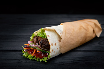 Shawarma rolled in lavash, moist grilled meat with onion, herbs and vegetables on wooden black background.