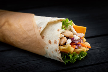 Shawarma rolled in lavash, moist grilled meat with onion, herbs and vegetables on wooden black background.