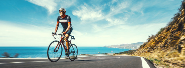 Naklejka na ściany i meble Mature Adult on a racing bike climbing the hill at mediterranean sea landscape coastal road