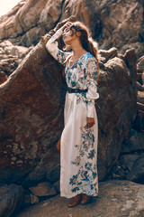 beautiful young woman in elegant stylish dress on stone beach at sunset