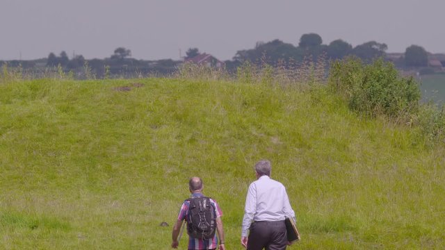 A point-of-view, medium-long shot of a couple of men walking up to the top of open grassy hills on a very sunny day.