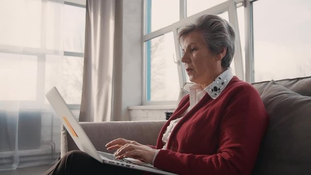 Happy american woman in her 50s scrolling through desktop interior agency cheerful, slowmo. Portrait of mature businesswoman session at desk work on laptop.