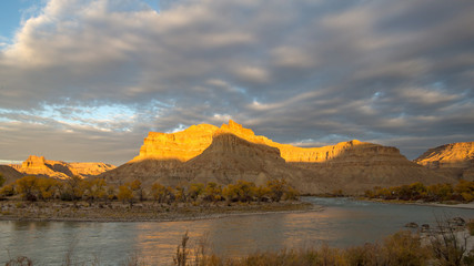 Sun lights up desert cliffs over the Green River with dark clouds in the sky in the Utah desert.