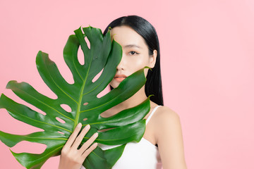 Portrait of young beautiful Asian woman holding tropical leaf on pink background. Natural beauty concept.