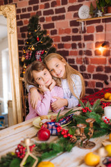 Fototapeta na wymiar Cute sisters in pajamas have fun in the kitchen by the table with gingerbread cookies, tangerines at home in the room decorated with Christmas garlands, needles. Christmas mood