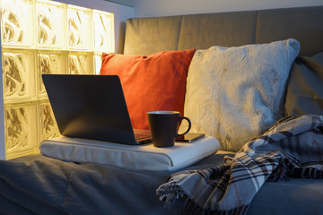 Laptop, phone, coffee cup, stand on gray sofa, plaid, pillows, glass backlight wall. Cosiness, work at home, online study concept. Close-up, copy space