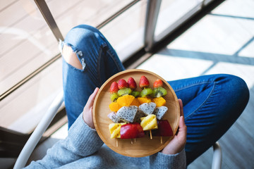 Top view image of a woman holding a wooden plate of fresh mixed fruits on skewers