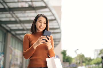 Portrait of elegant young Asian woman holding shopping bags and using smartphone on the go while...