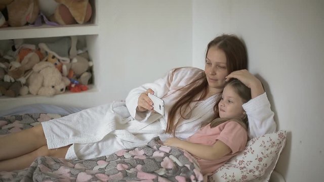 a little girl lies on the bed with her mother and watches cartoons on the smartphone screen, children and digital technology . Family relations of children and parents