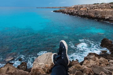 Schilderijen op glas Blurred men's feet in shoes on a background of sea and rocks. Relaxation by the sea. Landscapes of Cyprus © WellStock