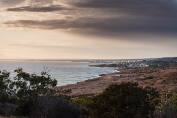 Long rocky shore with trees and restless sea. Houses in the background. Cape Greco, Cyprus