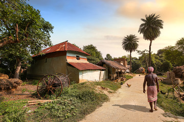 Rural man walking along an unpaved village road with view of mud houses at sunset. Photograph shot at Bolpur, West Bengal India - obrazy, fototapety, plakaty