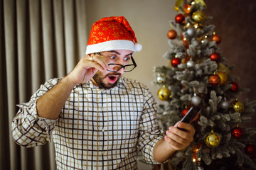 Portrait of a shocked man holding the phone in his hand, near a Christmas tree.