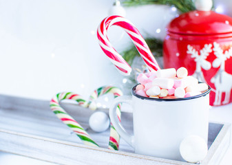 a white cup with marshmallows and Christmas sweets, on a white background with fir branches, balls and Christmas lights, as well as with a large red jar with deers in the background