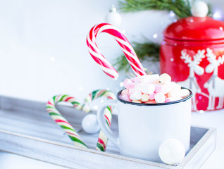 a white cup with marshmallows and Christmas sweets, on a white background with fir branches, balls and Christmas lights, as well as with a large red jar with deers in the background