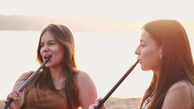 Two girls are smoking hookas and talking.The young are sitting at sunset. They are sitting at the lakeside.