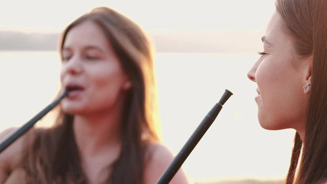 Two girls are smoking hookas and talking.The young are sitting at sunset. They are sitting at the lakeside.