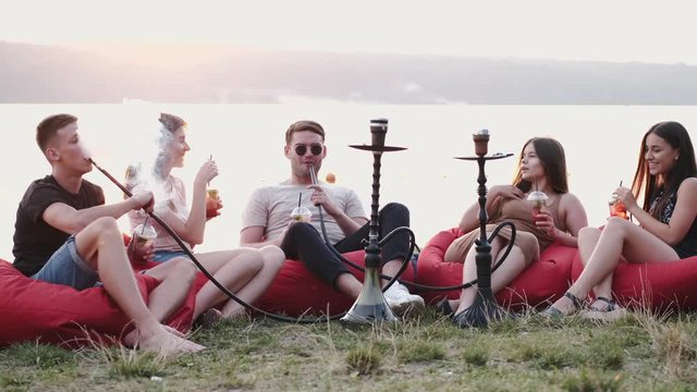 A circle of friends are smoking kalians .The young are sitting in the beanbag chairs at sunset. They are sitting at the lakeside.