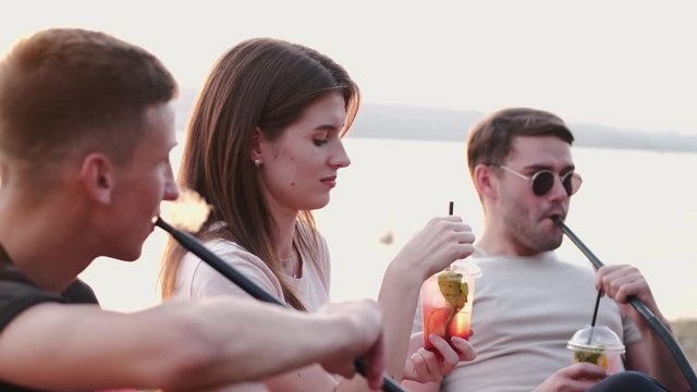 Young people are vaping hookahs and drinking cocktail. The boys are smoking and the girl is drinking a cocktail. They are sitting at sunset at the beach.