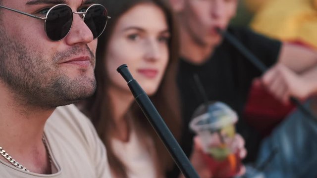Two European boys are smoking and a girl is drinking a cocktail. The girl is smiling. The boy is wearing sunglasses. They are chilling at sunset.