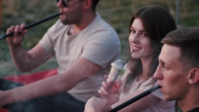 Two European boys are smoking and a girl is drinking a cocktail. The girl is smiling. They are chilling at sunset.