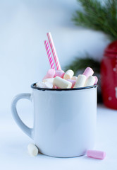 a white cup with marshmallows on a white background with fir branches, balls and Christmas lights, as well as a large red jar with deers in the background