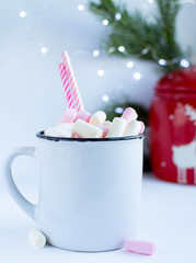a white cup with marshmallows on a white background with fir branches, balls and Christmas lights, as well as a large red jar with deers in the background