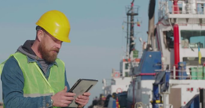 Port Worker With A Beard In A Yellow Helmet Stands With A Tablet PC In The Seaport Against The Background Of Ship. The Docker Inspects The Industrial Harbor. Cinema 4K Video