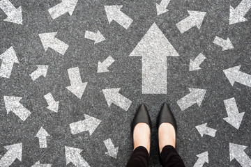 Feet and arrows on road background from above. Businesswoman standing on pathway with drawn white...