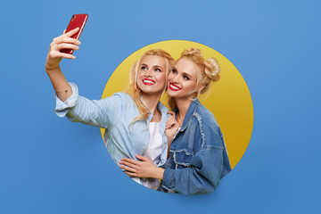 Image of two young happy girlfriends women standing over a blue background. Twins peep out of a hole in the wall. Looking to the side to take a selfie on the mobile phone.