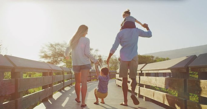 Happy parents walking and swinging their young toddler son into the air, young toddler son riding on dads shoulders, family fun on a coastal boardwalk at sunset, weekend family walk
