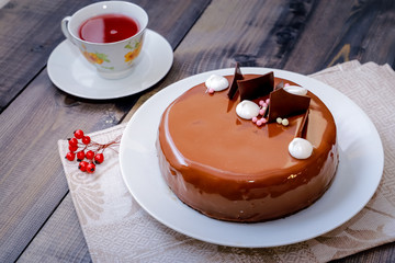 On a dark tabletop, on a linen napkin on a white dish, there is a round chocolate cake, next to a white cup is dark tea and next to the round berries