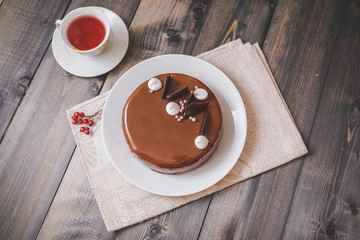 On a dark tabletop, on a linen napkin on a white dish, there is a round chocolate cake, next to a white cup is dark tea and next to the round berries