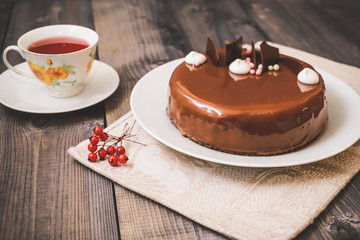 On a dark tabletop, on a linen napkin on a white dish, there is a round chocolate cake, next to a white cup is dark tea and next to the round berries