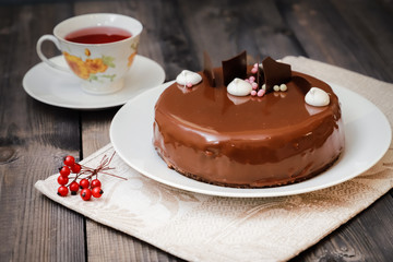 On a dark tabletop, on a linen napkin on a white dish, there is a round chocolate cake, next to a white cup is dark tea and next to the round berries